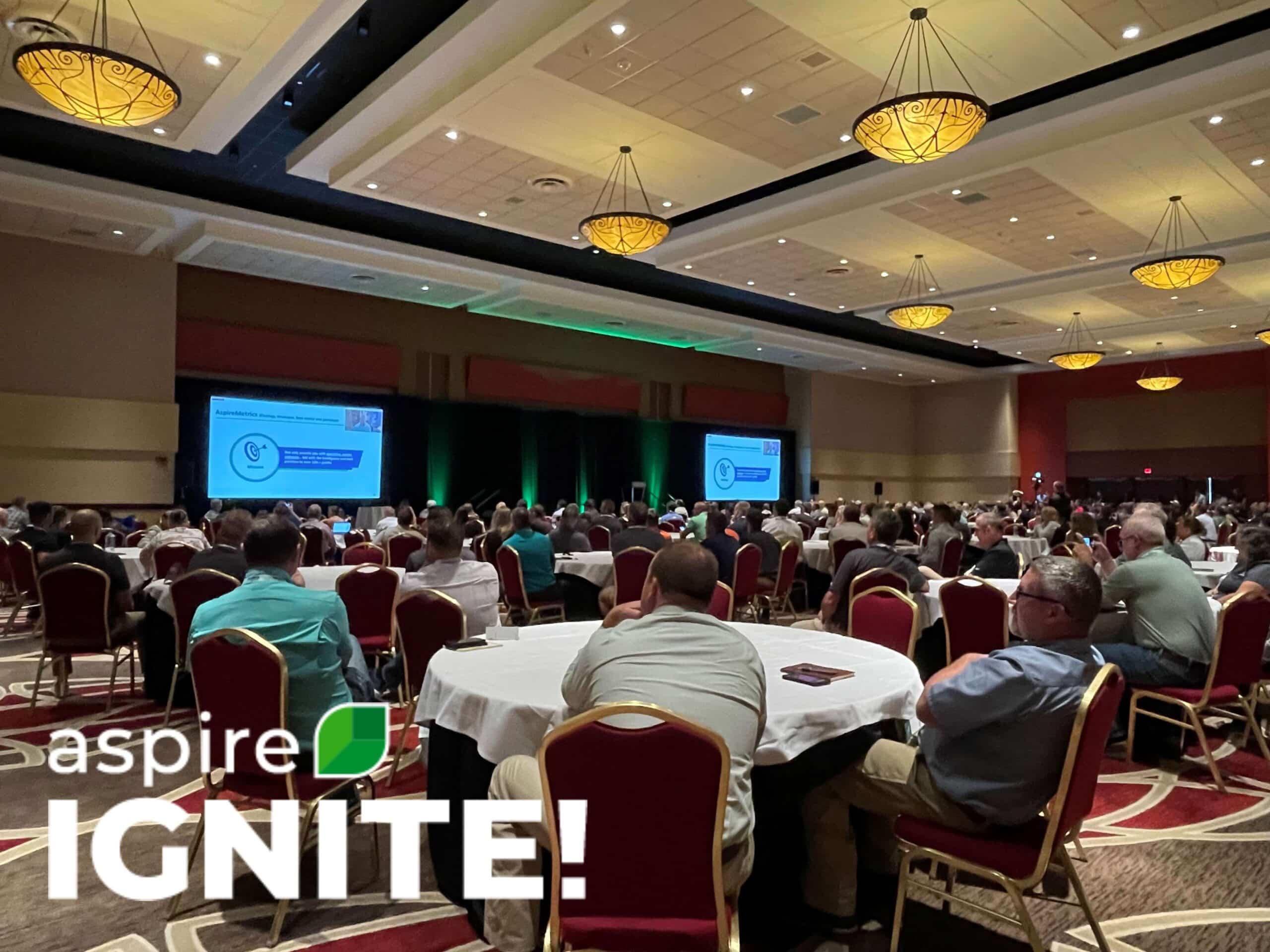 Aspire IGNITE event with attendees seated at round tables in a large ballroom with presentations showcased on screens