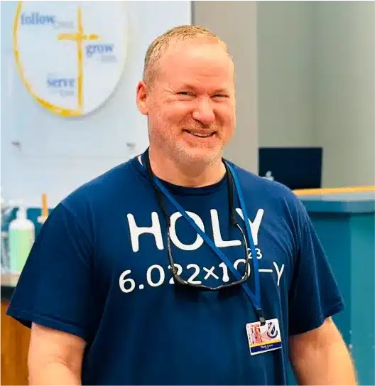 Smiling staff member wearing a chemistry-themed t-shirt in a classroom with a follow, grow, serve wall graphic