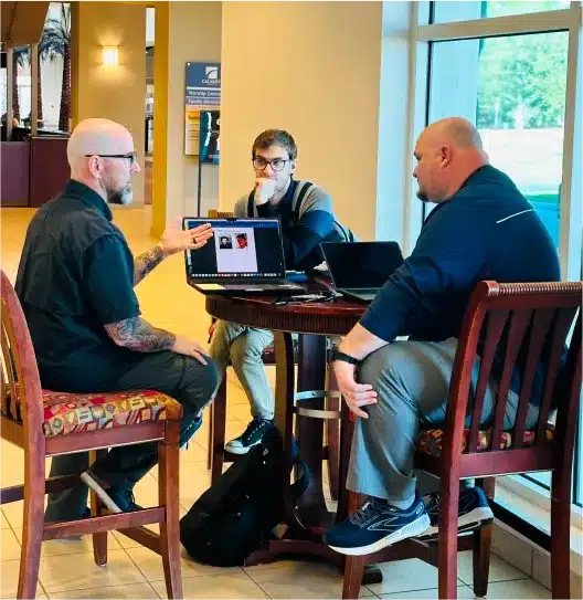 Three men discussing information on laptops at a small table in an office lobby