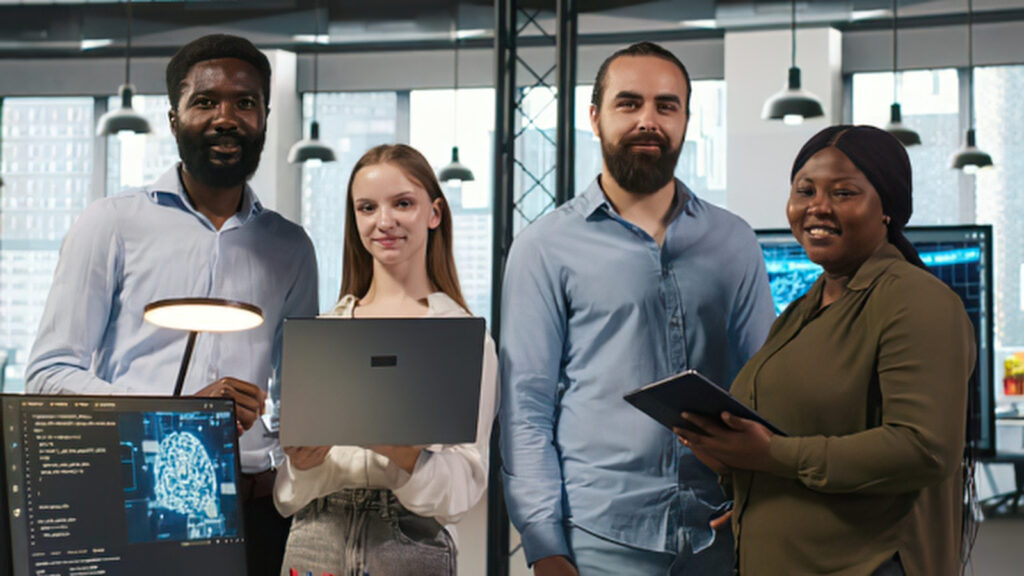 Four IT professionals smiling together in an office with monitors displaying code