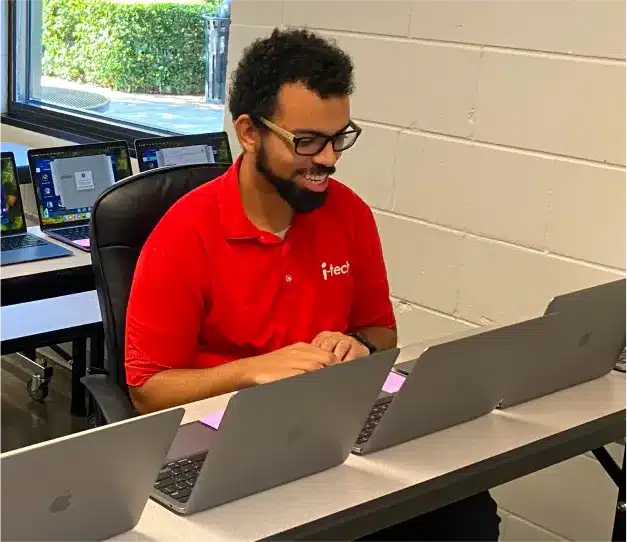 i-Tech technician smiling while working on a laptop among a row of laptops in a classroom