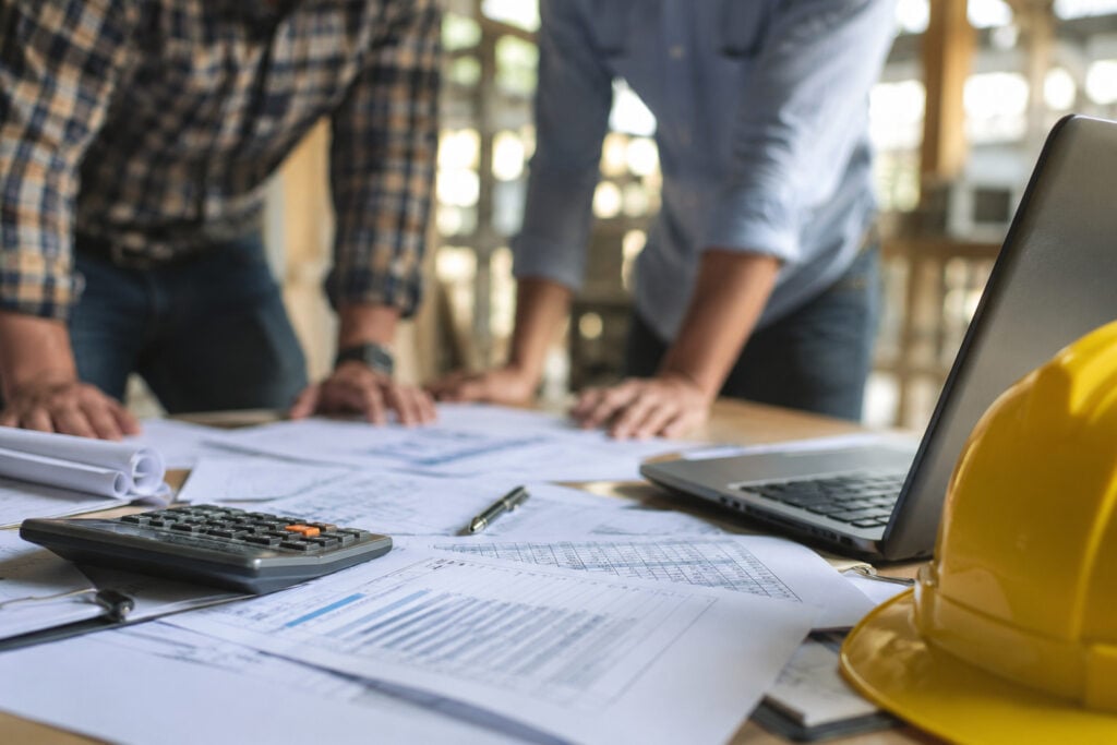 Calculator, job costing spreadsheets, blueprints, and a yellow hard hat on a construction table